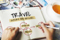 Person writing travel plans in notebook with vacation and holiday ideas, tea cup and photos on desk