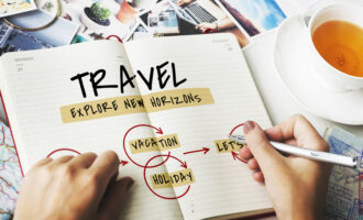 Person writing travel plans in notebook with vacation and holiday ideas, tea cup and photos on desk